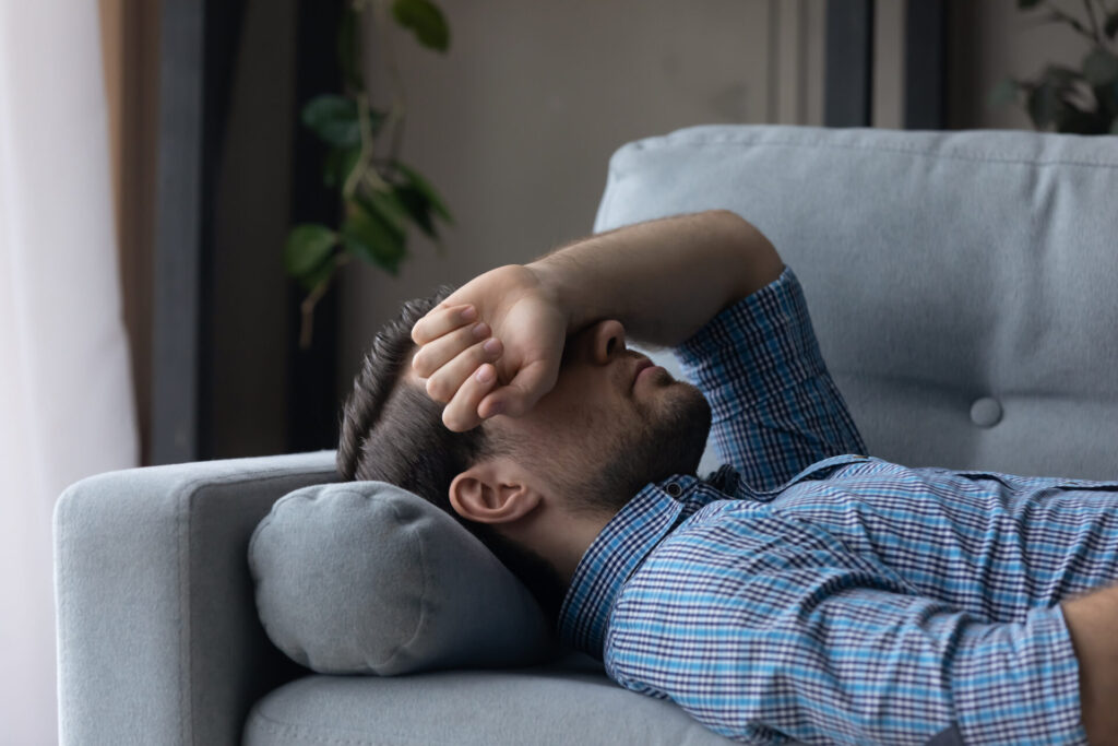 Sick man feeling headache migraine or dizziness, covering eyes with hand, lying on couch at home, exhausted tired millennial male sleeping after difficult working day, resting on comfortable sofa